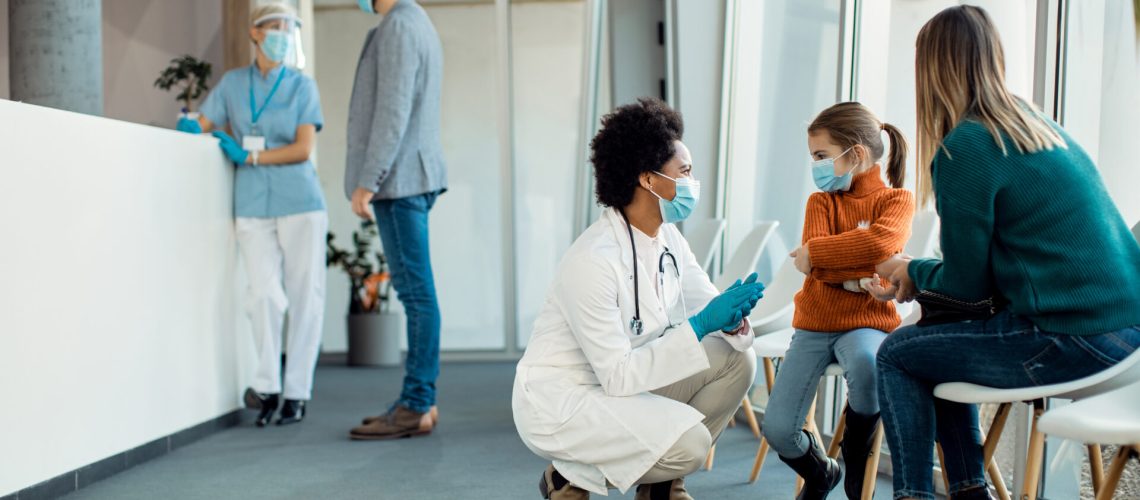 Little girl pouting while African American female doctor is talking to her in a waiting room at the hospital. They are wearing protective face mask dur to coronavirus epidemic.
